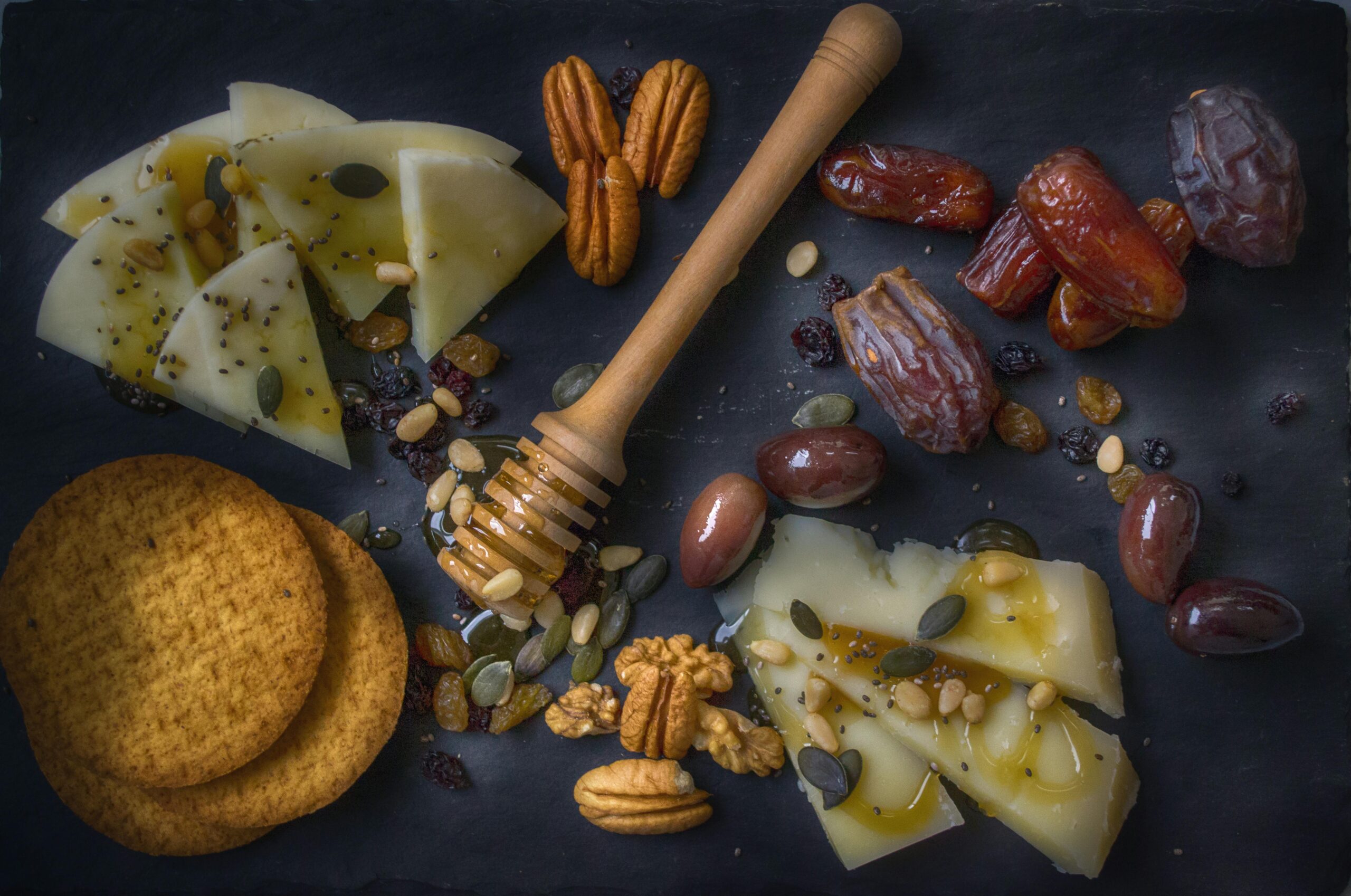 A delightful snack assortment with cheese slices, nuts, dates, and honey on a slate board.